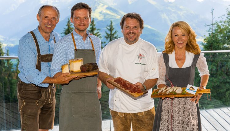 Vier lächelnde Menschen in traditioneller österreichischer Alpentracht halten Teller mit Essen, während im Hintergrund die Berge zu sehen sind.