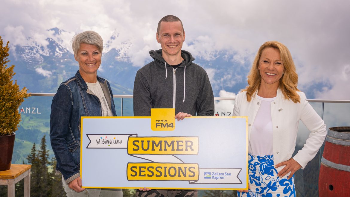 Drei Personen lächeln und halten ein Summer Sessions Schild mit Bergen und bewölktem Himmel im Hintergrund.