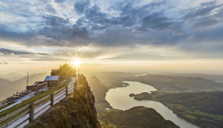 Die Sonne geht über einer Berghütte auf, mit Blick auf einen See und sanfte Hügel unter einem dramatischen, bewölkten Himmel.