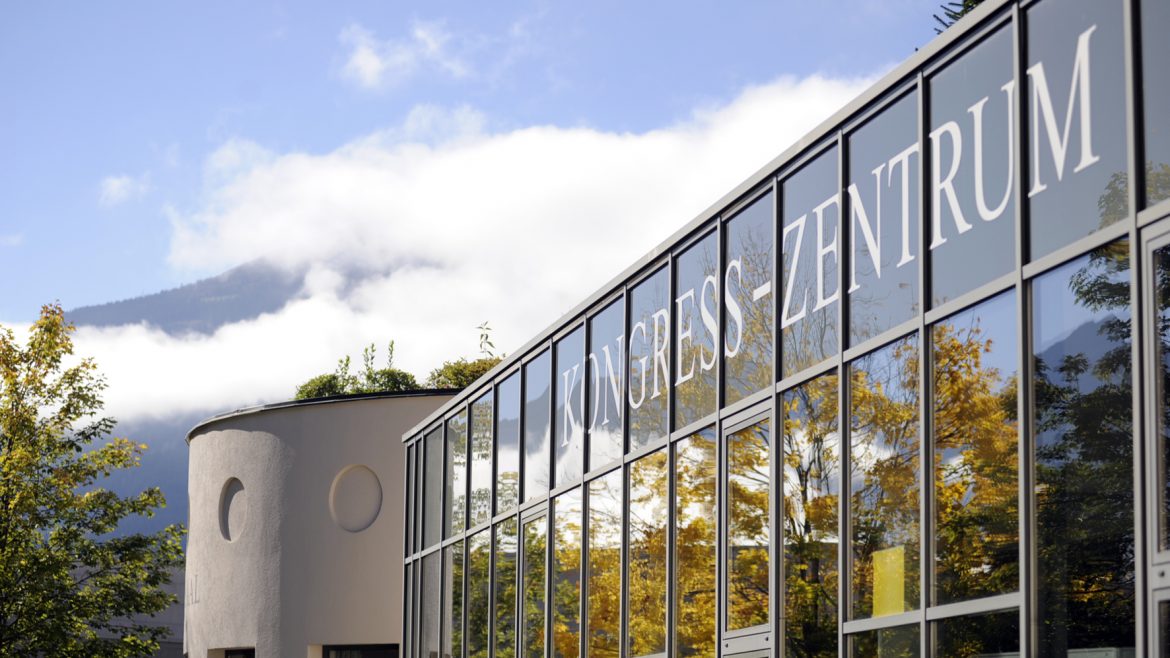 Modernes Glasgebäude mit der Aufschrift Kongress-Zentrum mit Bäumen und Bergen im Hintergrund.
