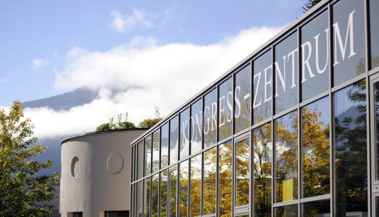 Modernes Glasgebäude mit der Aufschrift Kongress-Zentrum mit Bäumen und Bergen im Hintergrund.