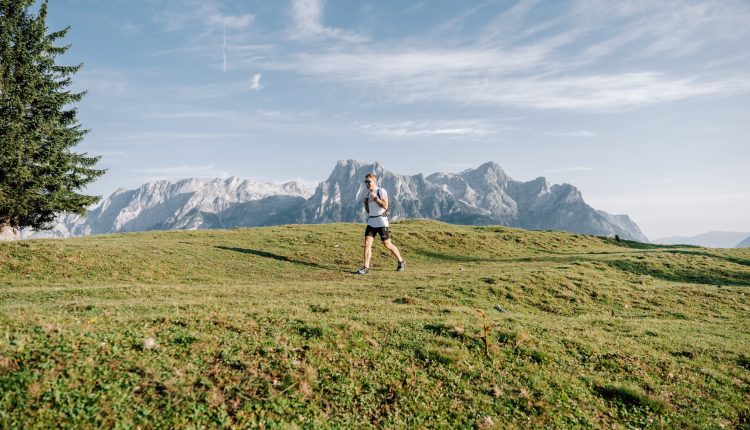 Person beim Wandern auf einem grasbewachsenen Hügel mit Bergen und blauem Himmel im Hintergrund.