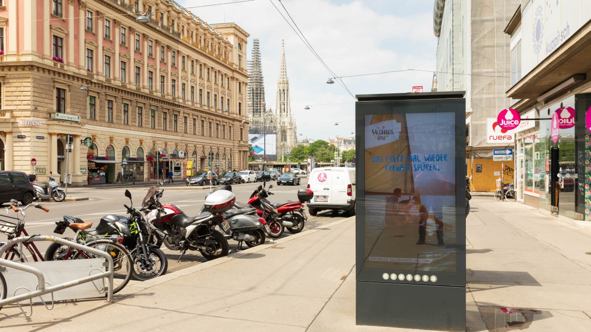 Eine Stadtstraße mit geparkten Motorrädern, Autos und einer digitalen Werbetafel; im Hintergrund historische Gebäude.