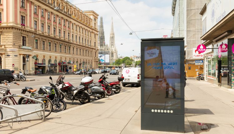Eine Stadtstraße mit geparkten Motorrädern, Autos und einer digitalen Werbetafel; im Hintergrund historische Gebäude.