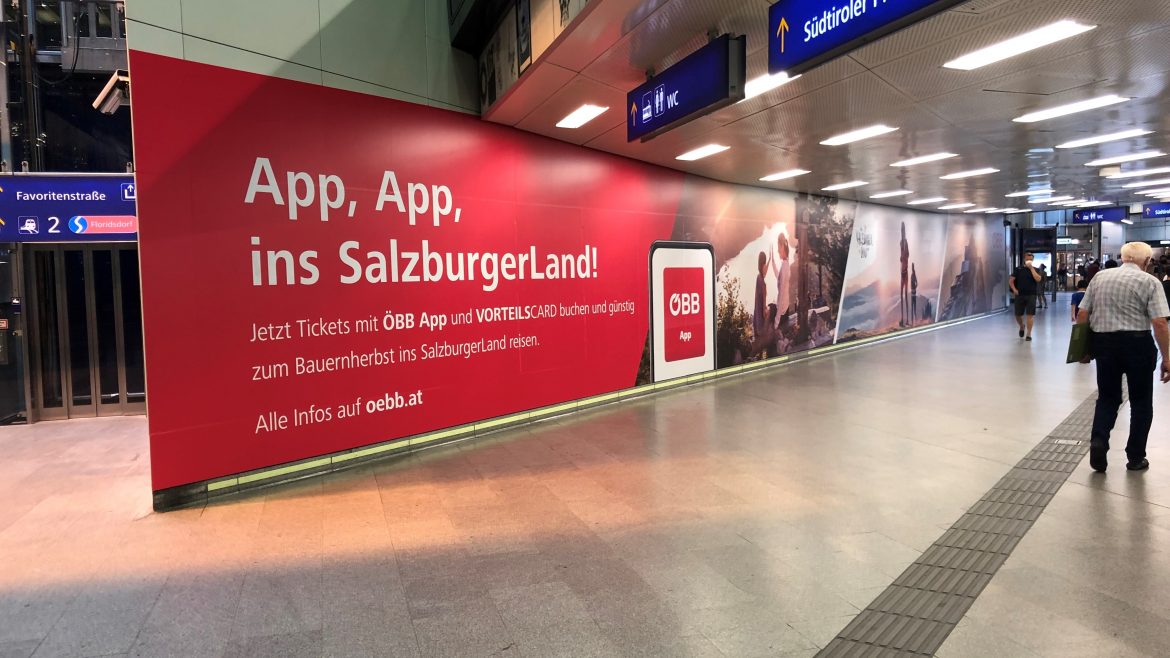 Große rote Werbung in einem Bahnhof, die für die ÖBB-App im SalzburgerLand wirbt, mit vorbeilaufenden Reisenden.