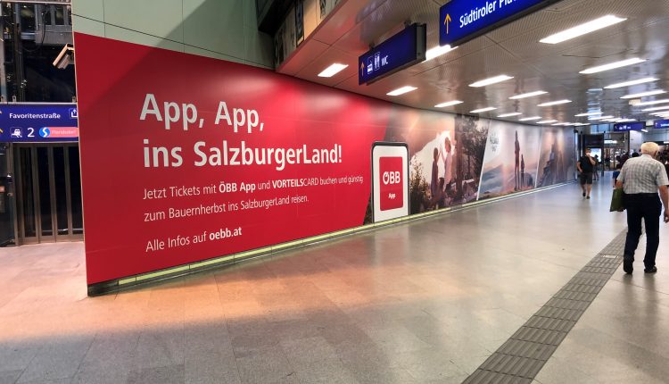 Große rote Werbung in einem Bahnhof, die für die ÖBB-App im SalzburgerLand wirbt, mit vorbeilaufenden Reisenden.