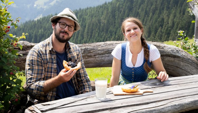 Ein Mann und eine Frau sitzen an einem rustikalen Tisch im Freien und genießen Gebäck und Milch mit einem malerischen Bergblick im Rücken.