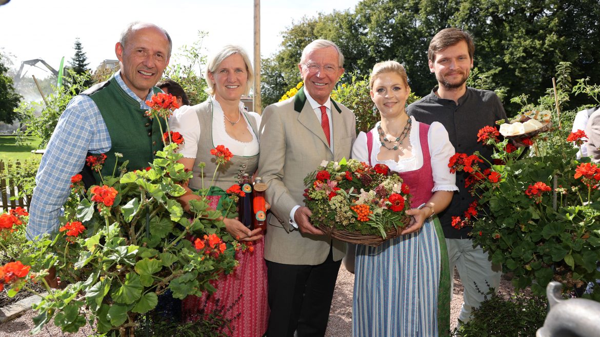 Fünf Personen in österreichischer Tracht stehen im Freien zwischen Blumen, lächeln und halten einen Korb mit Blüten.