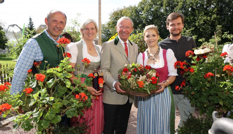 Fünf Personen in österreichischer Tracht stehen im Freien zwischen Blumen, lächeln und halten einen Korb mit Blüten.