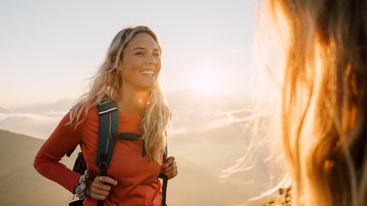 Lächelnde Frau mit einem Rucksack wandert bei Sonnenaufgang, Berge und Sonnenlicht im Hintergrund.