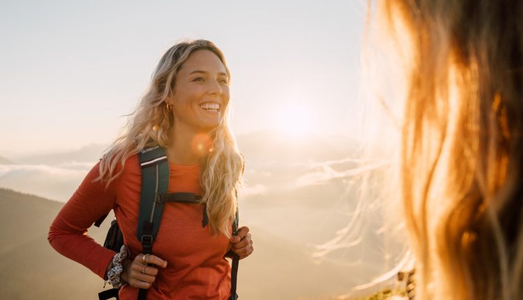 Lächelnde Frau mit einem Rucksack wandert bei Sonnenaufgang, Berge und Sonnenlicht im Hintergrund.