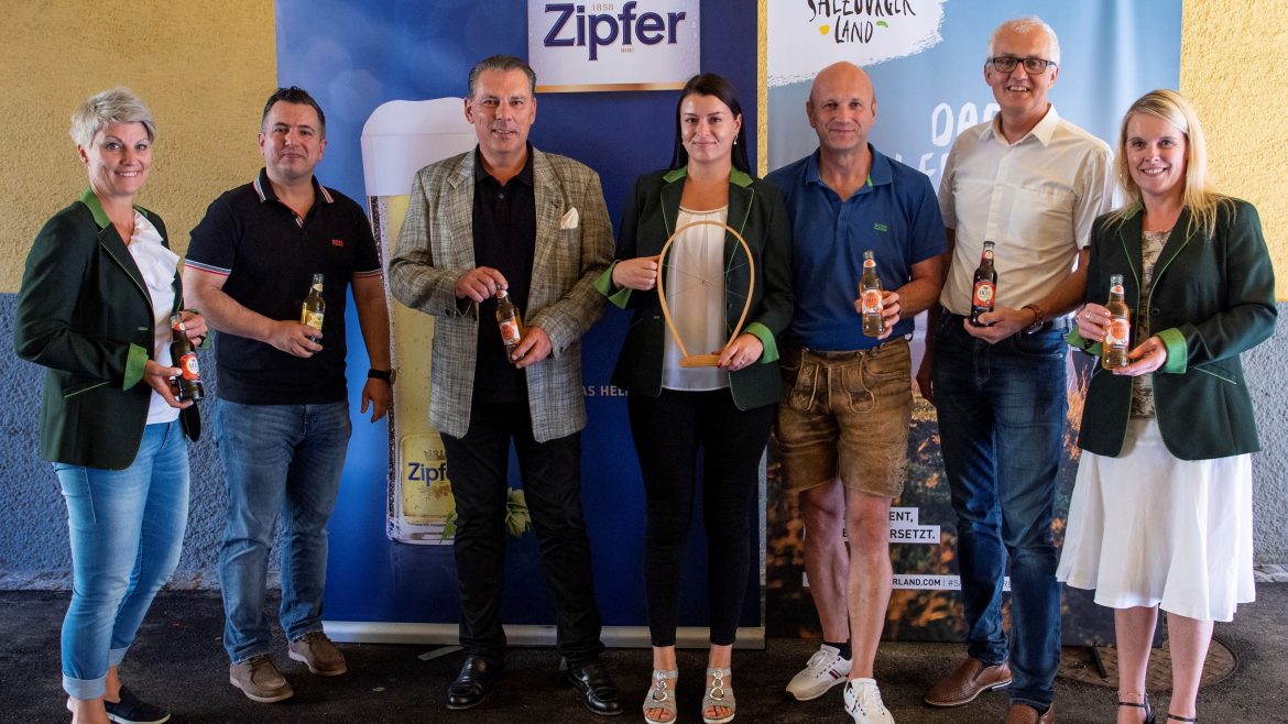 Sieben Personen stehen lächelnd und mit Bierflaschen in der Hand vor Zipfer- und Salzburger Land-Transparenten.