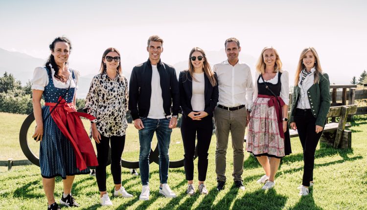Sieben Menschen stehen lächelnd im Freien auf einer Wiese, mit Bergen im Hintergrund, einige in traditioneller österreichischer Kleidung.