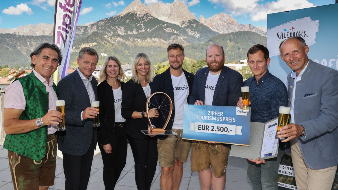 Acht Personen, die mit Getränken lächeln, eine Trophäe und einen Scheck in der Hand halten, mit Bergen im Hintergrund.