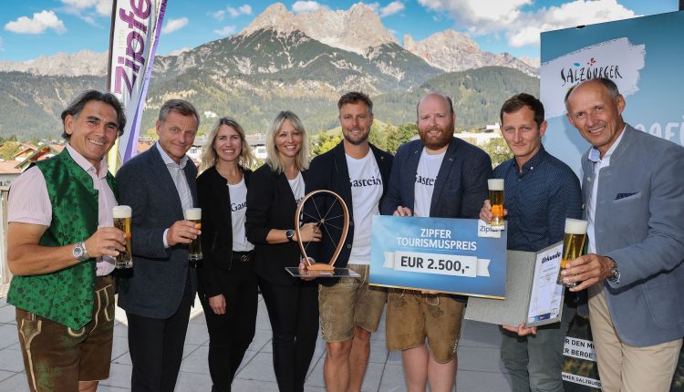 Acht Personen, die mit Getränken lächeln, eine Trophäe und einen Scheck in der Hand halten, mit Bergen im Hintergrund.