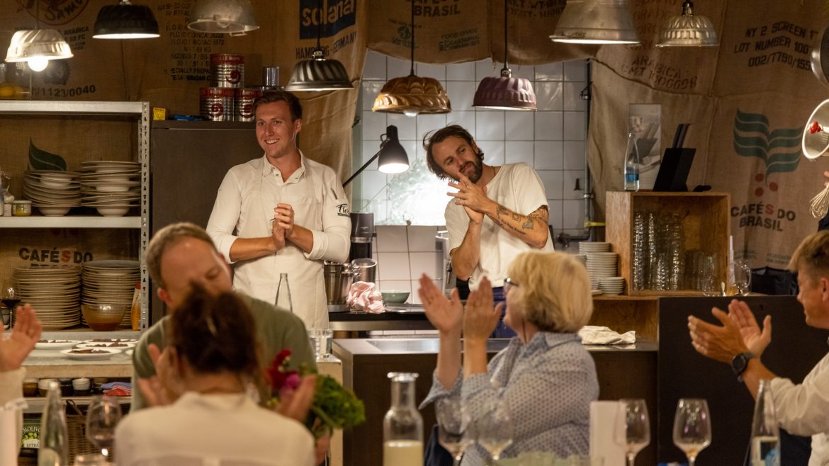 Zwei Köche stehen in einem warm beleuchteten, rustikalen Restaurant und lächeln, während die Gäste ihnen applaudieren.