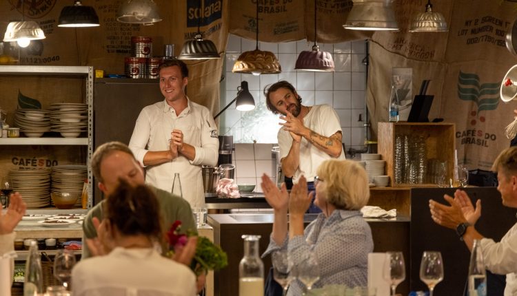 Zwei Köche stehen in einem warm beleuchteten, rustikalen Restaurant und lächeln, während die Gäste ihnen applaudieren.