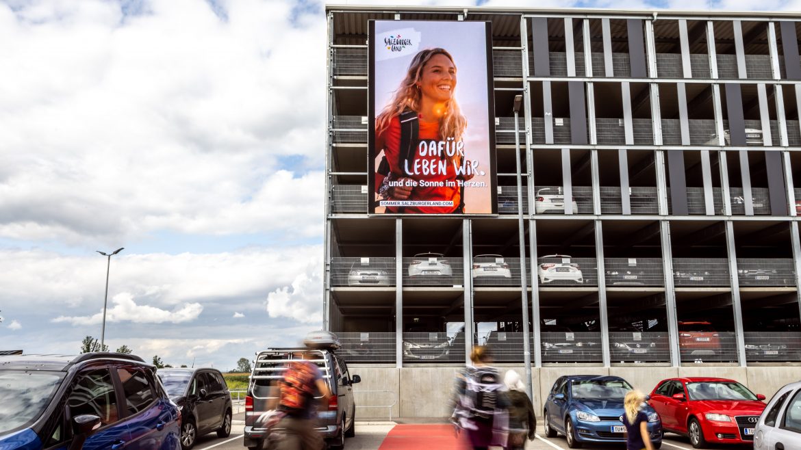 Menschen gehen in der Nähe eines Parkhauses vorbei, an dessen Fassade eine große Werbung mit einer lächelnden Frau angebracht ist.