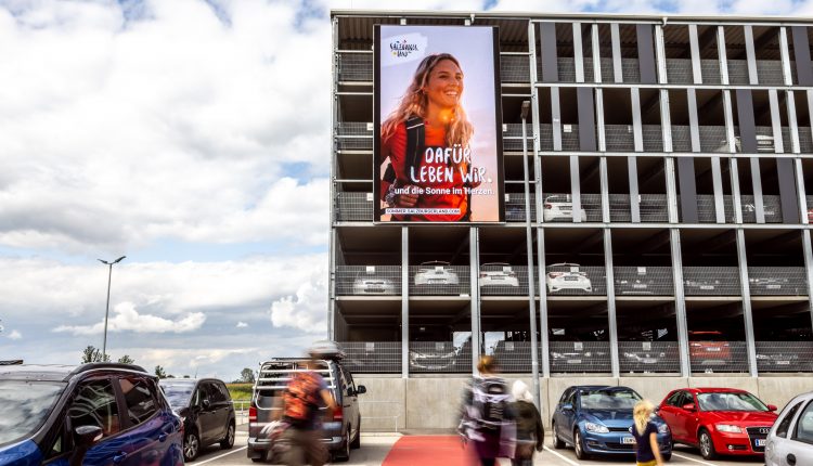 Menschen gehen in der Nähe eines Parkhauses vorbei, an dessen Fassade eine große Werbung mit einer lächelnden Frau angebracht ist.