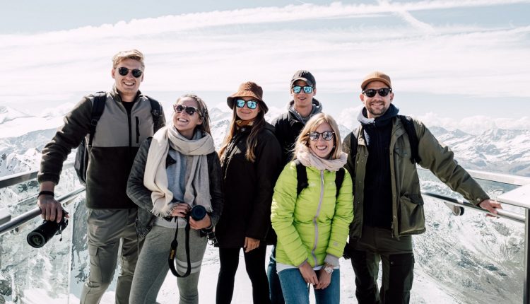 Sechs Personen in Winterkleidung posieren auf einer verschneiten Aussichtsplattform in den Bergen mit blauem Himmel und Wolken im Hintergrund.