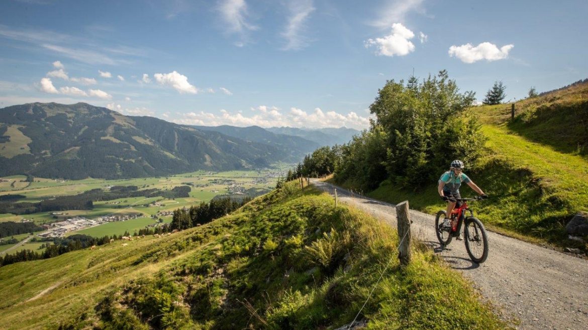 Eine Person fährt mit dem Mountainbike auf einem Weg mit Blick auf ein malerisches Tal mit Bergen im Hintergrund.