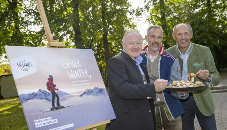 Drei Männer in Jacken stehen im Freien neben einem Poster eines Skifahrers, lächeln und halten einen Teller mit Essen in der Hand.