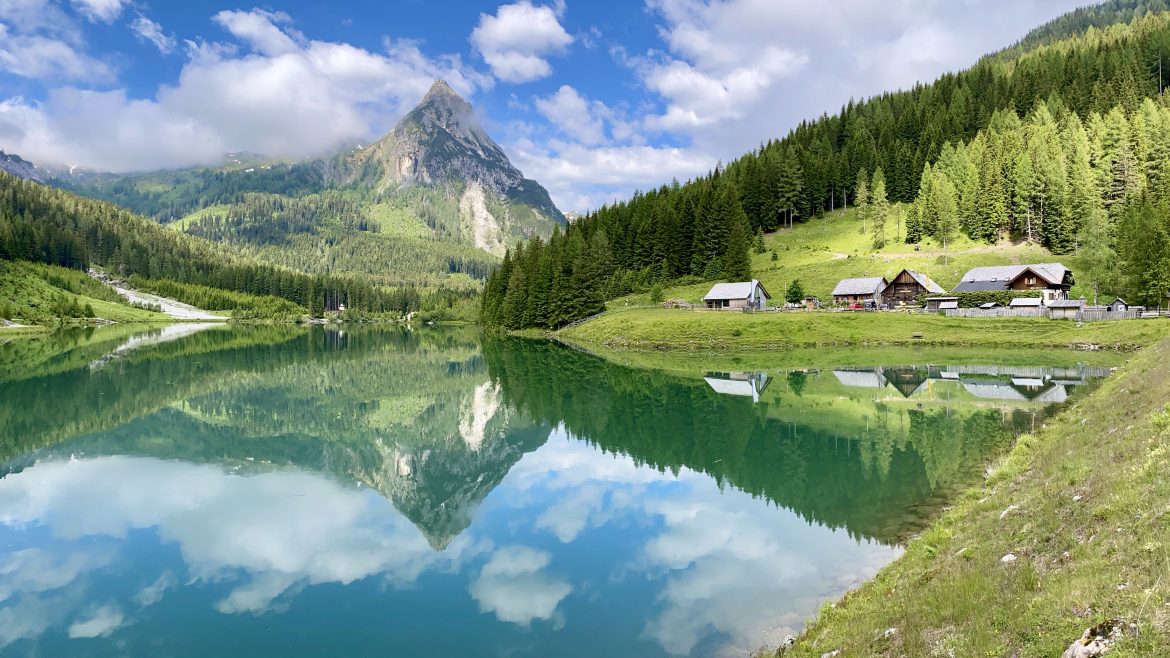 In einem ruhigen See spiegeln sich Berge, immergrüne Bäume und Hütten unter einem teilweise bewölkten blauen Himmel.