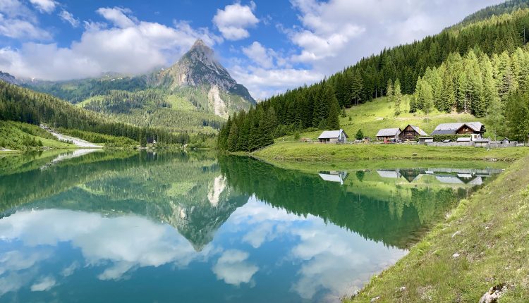 In einem ruhigen See spiegeln sich Berge, immergrüne Bäume und Hütten unter einem teilweise bewölkten blauen Himmel.