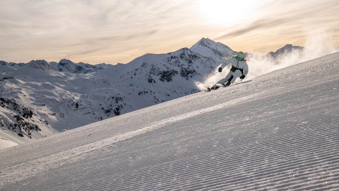 Ein Skifahrer fährt auf einer präparierten Schneepiste mit Bergen und einem bewölkten Himmel im Hintergrund.