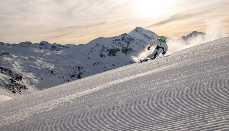 Ein Skifahrer fährt auf einer präparierten Schneepiste mit Bergen und einem bewölkten Himmel im Hintergrund.