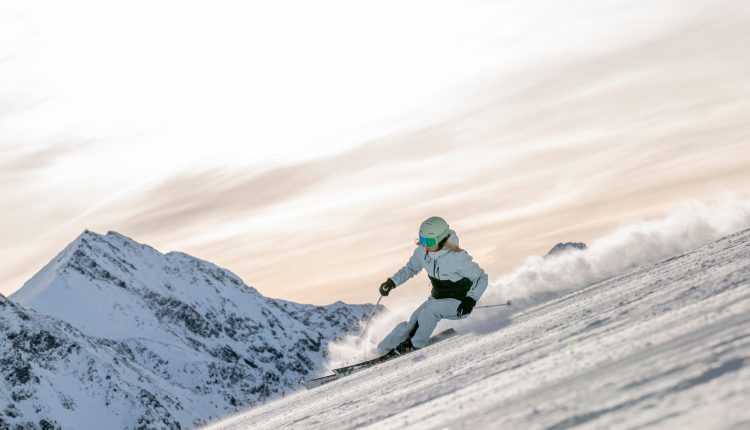 Ein Skifahrer in weißer Kleidung fährt auf einem verschneiten Berghang mit Gipfeln im Hintergrund bergab.