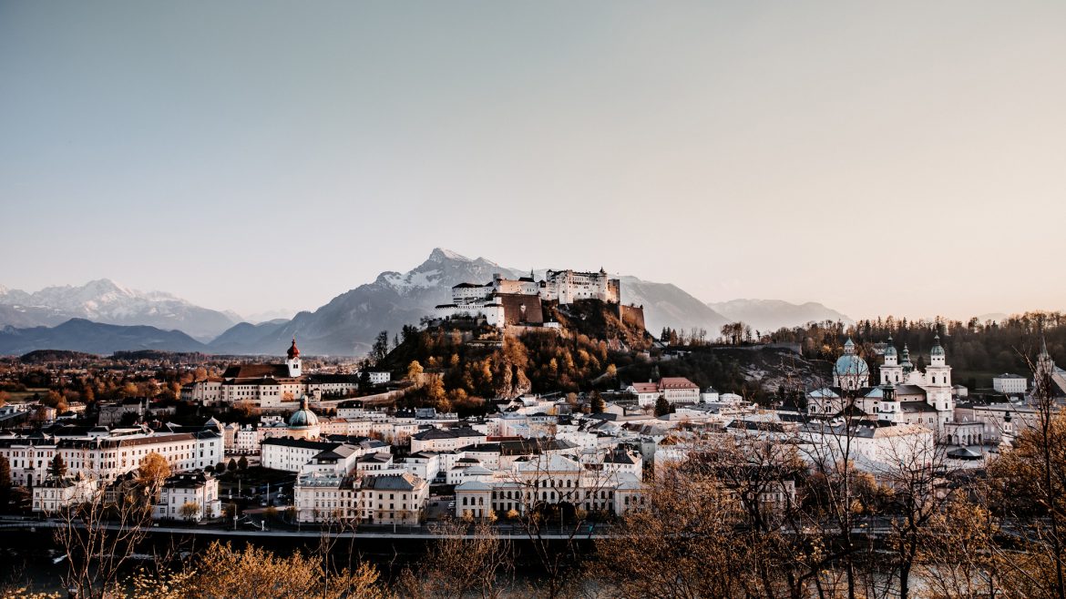 Ansicht von Salzburg, Österreich, mit der Festung Hohensalzburg auf einem Hügel und Bergen im Hintergrund bei Sonnenuntergang.