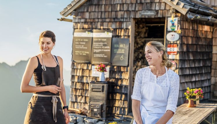 Zwei Frauen in Schürzen lächeln vor einem rustikalen Cafe-dafür leben wir, für Momente wie diese in den Bergen.