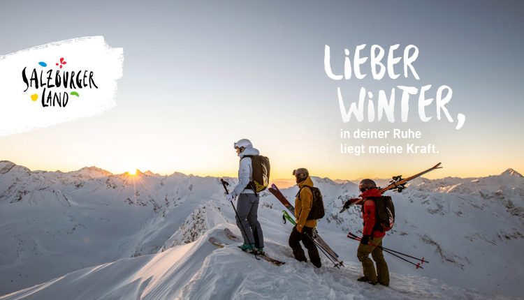 Drei Skifahrer stehen bei Sonnenaufgang auf einem verschneiten Berggipfel und bewundern die Winterlandschaft.