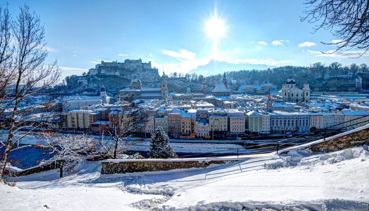 Schneebedecktes Stadtbild von Salzburg, Österreich, mit einer strahlenden Sonne und einer Festung auf einem Hügel im Hintergrund.