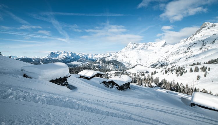 Schneebedeckte Hütten an einem Hang mit hohen Bergen und blauem Himmel im Hintergrund.
