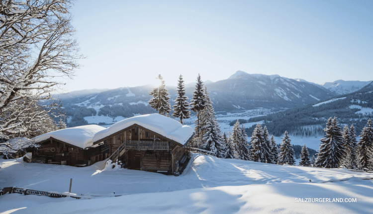 Schneebedeckte Hütten und Kiefern an einem Berghang mit fernen Gipfeln unter einem klaren blauen Himmel.