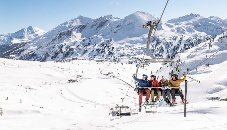 Vier Skifahrer fahren an einem hellen, klaren Tag mit einem Skilift über verschneite Berge.