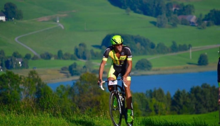 Radfahrer mit gelbem Helm fährt bergauf auf einer Landstraße mit grünen Hügeln und einem See im Hintergrund.