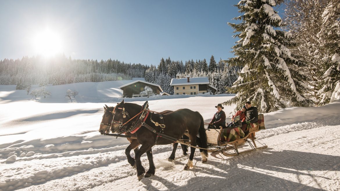 Zwei Personen fahren in einem Pferdeschlitten durch eine verschneite Landschaft mit Bäumen und Häusern bei strahlendem Sonnenschein.