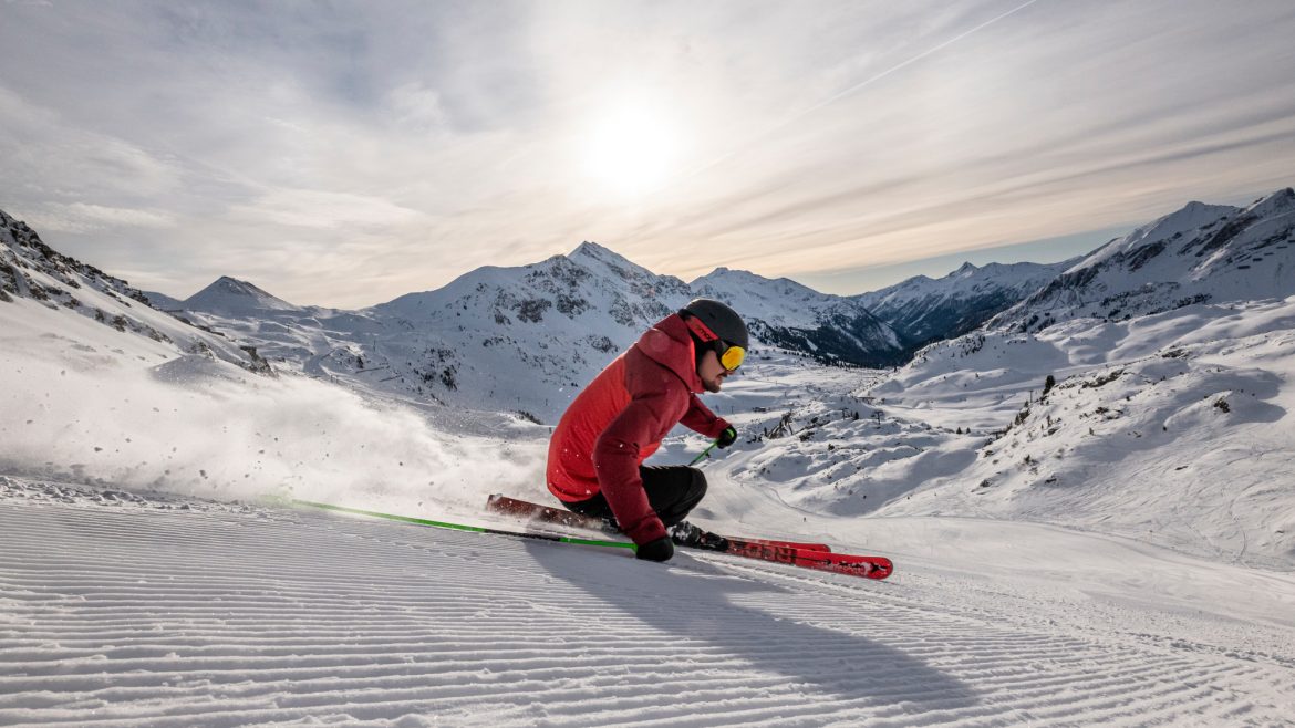 Skifahrer in einer roten Jacke, der bei Sonnenaufgang einen verschneiten Berghang hinunterfährt, mit Bergen im Hintergrund.