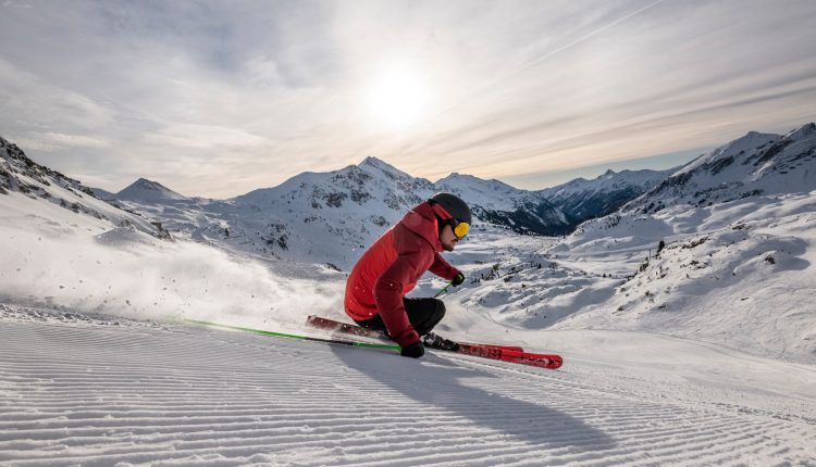 Skifahrer in einer roten Jacke, der bei Sonnenaufgang einen verschneiten Berghang hinunterfährt, mit Bergen im Hintergrund.