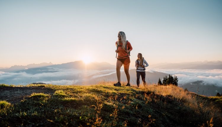 Zwei Frauen wandern auf einem grasbewachsenen Hügel bei Sonnenaufgang, mit Bergen und Wolken im Hintergrund.