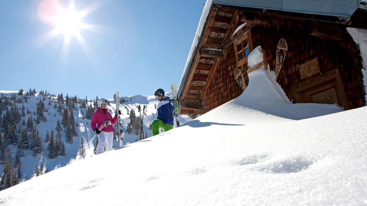 Zwei Skifahrer stehen an einem sonnigen Tag in den Bergen vor einer verschneiten Holzhütte, die Bäume und den blauen Himmel im Blick.