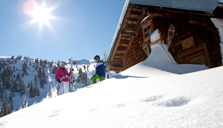 Zwei Skifahrer stehen an einem sonnigen Tag in den Bergen vor einer verschneiten Holzhütte, die Bäume und den blauen Himmel im Blick.