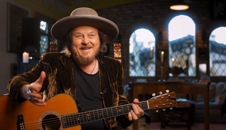 Ein lächelnder Mann mit Hut spielt eine Akustikgitarre in einem Innenraum, mit gewölbten Fenstern im Hintergrund.
