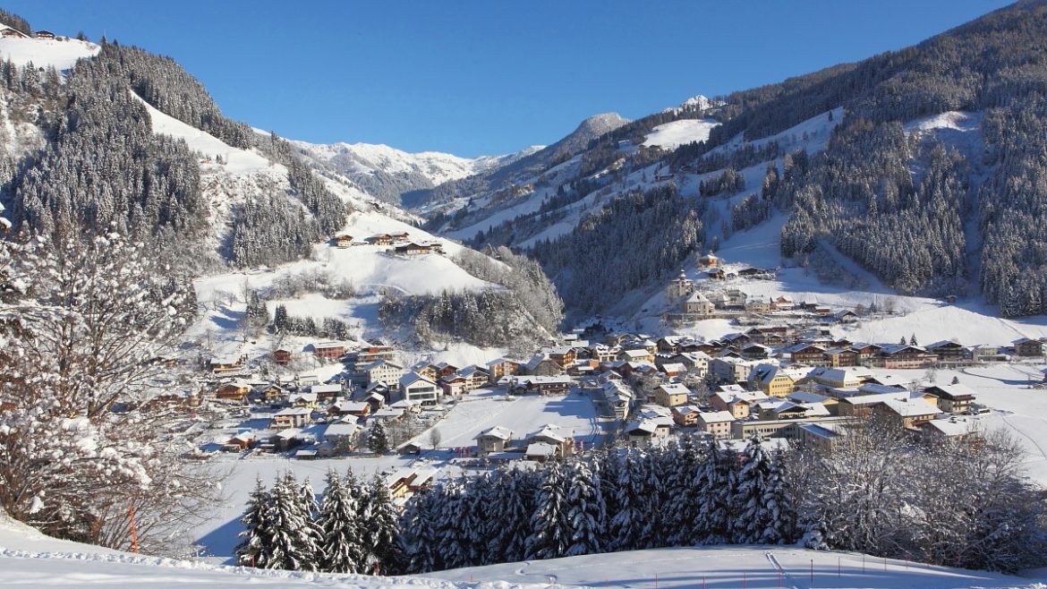 Ein verschneites Bergdorf mit Häusern inmitten von Kiefern unter einem strahlend blauen Himmel.