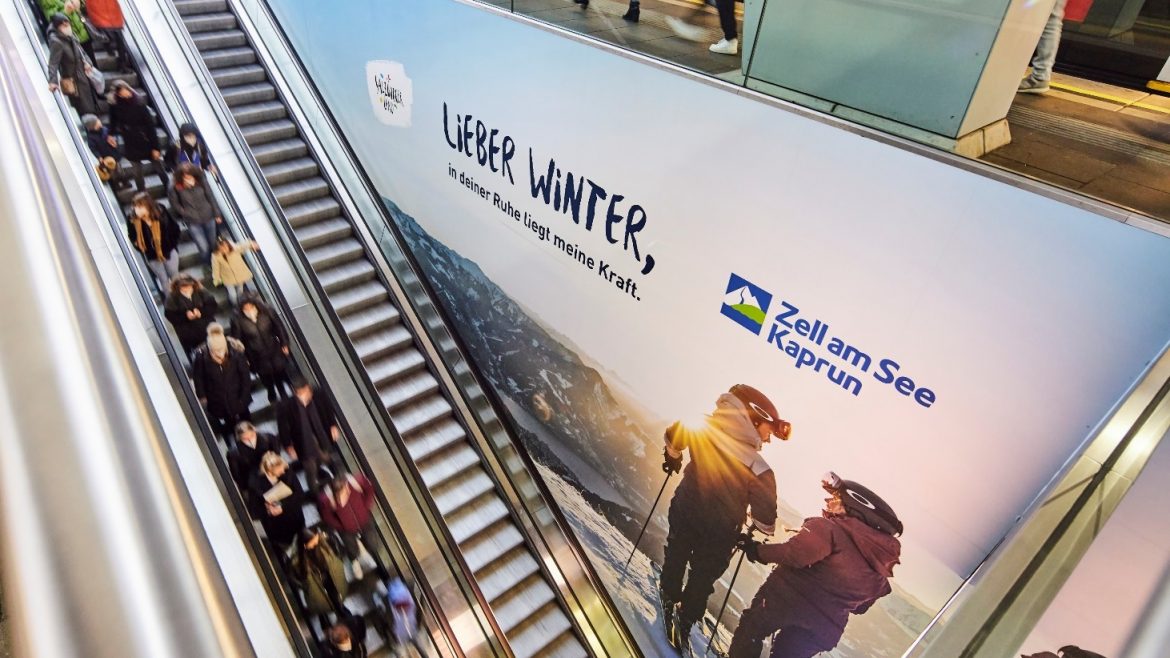 Menschen auf einer Rolltreppe neben einer großen Winterreisewerbung für Zell am See-Kaprun.