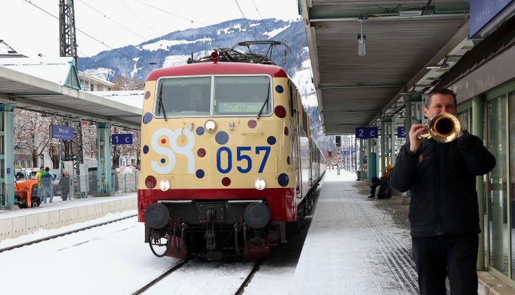 Ein Mann spielt auf einem verschneiten Bahnsteig Trompete, während ein Zug in den Bahnhof einfährt.
