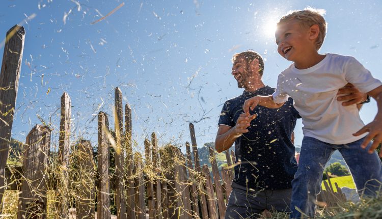 Ein Mann und ein Kind lachen zusammen, während sie an einem sonnigen Tag in der Nähe eines Holzzauns mit Stroh spielen.